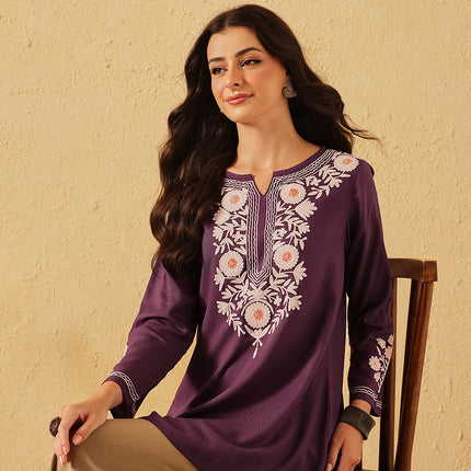 Woman wearing a purple embroidered dress sitting on a chair against a beige wall