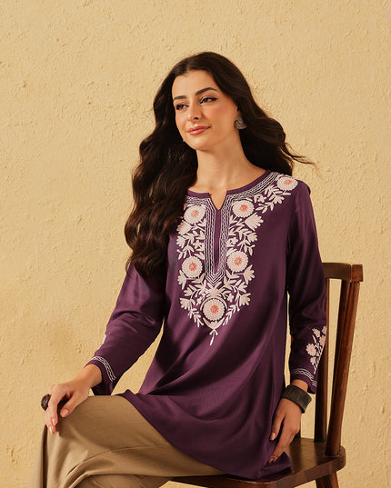 Woman wearing a purple embroidered dress sitting on a chair against a beige wall