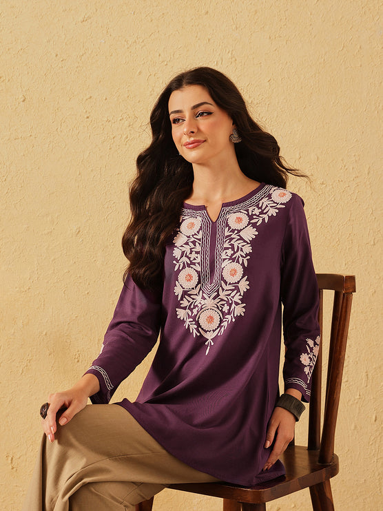 Woman wearing a purple embroidered dress sitting on a chair against a beige wall